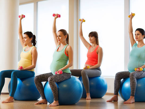 pregnancy, sport, fitness, people and healthy lifestyle concept - group of happy pregnant women with dumbbells exercising on ball in gym